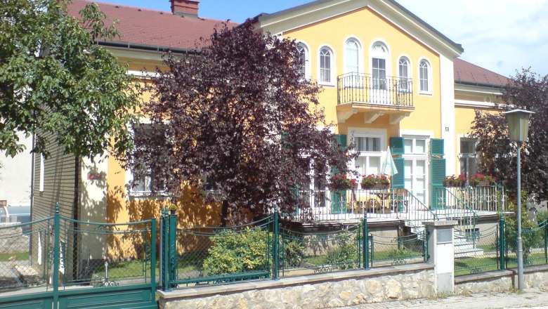 Gelbes Haus mit Balkon und grünem Zaun, umgeben von Bäumen.